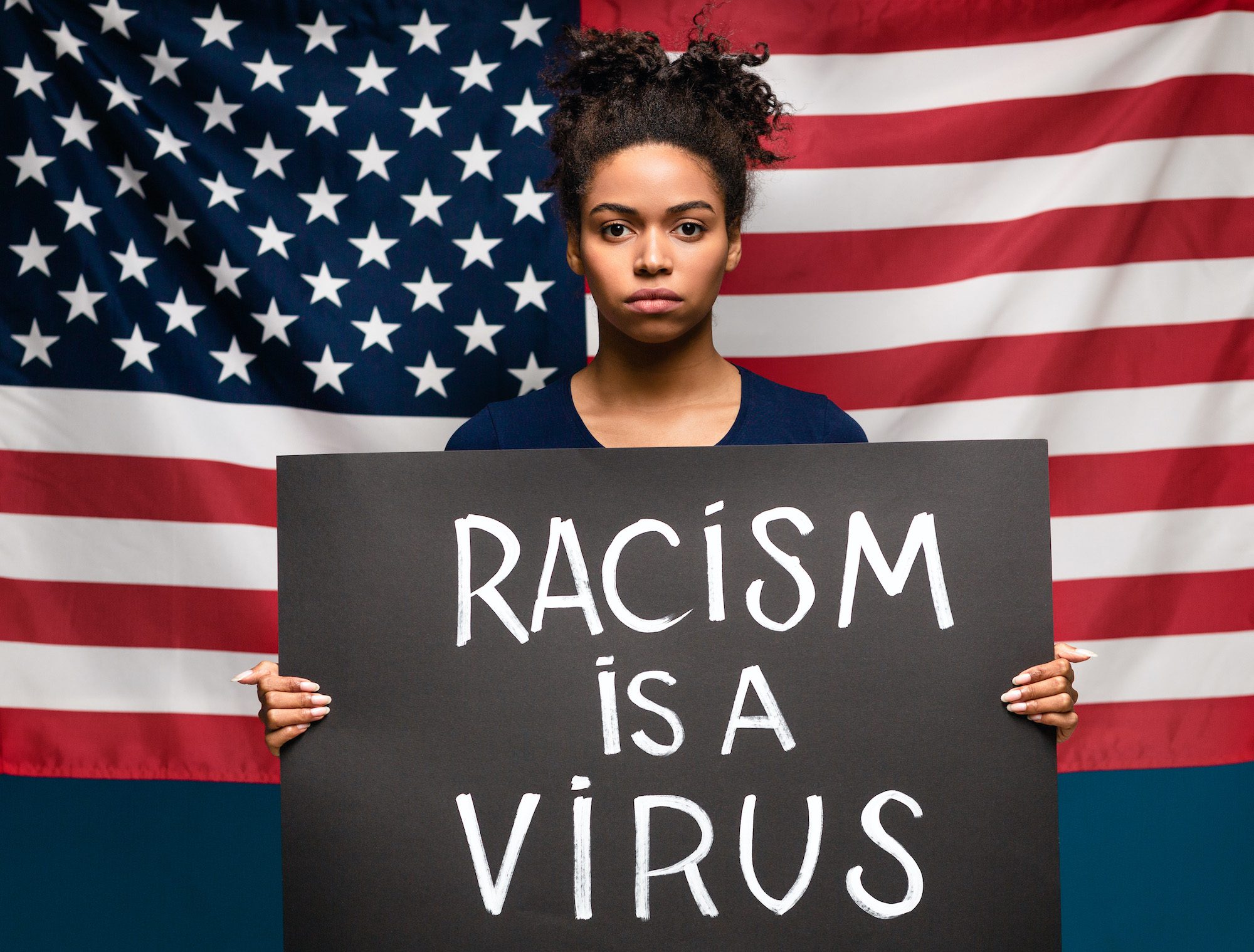 African American teenager standing in front of a flag and holding a placard that says "Racism is a virus."