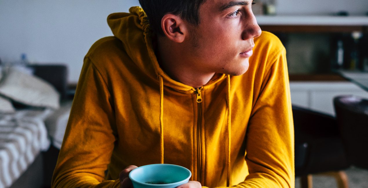 young adult college student looking sadly out bedroom window while drinking coffee