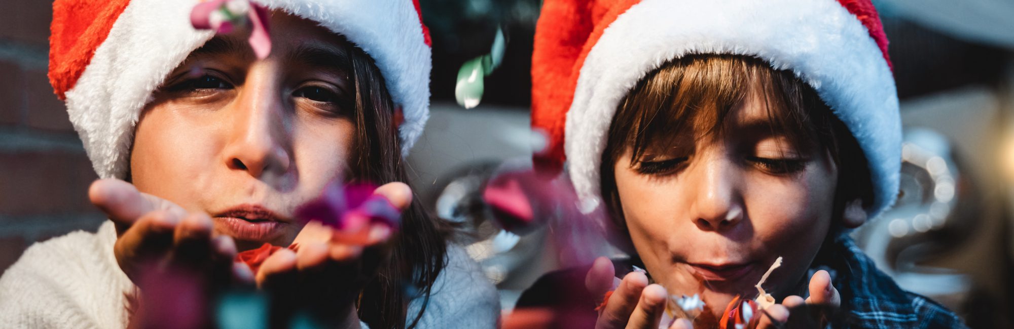 Holidays - Little girl and boy wearing Santa hats, blowing confetti from their hands towards camera