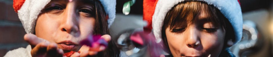 Holiday Stress - Little girl and boy wearing Santa hats, blowing confetti from their hands towards camera