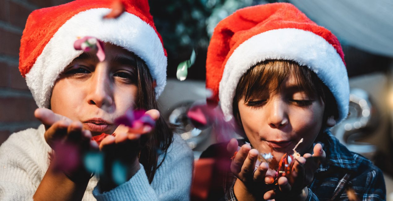 Holiday Stress - Little girl and boy wearing Santa hats, blowing confetti from their hands towards camera