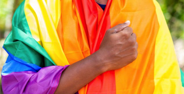 Photo of Man praying with clenched fist over heart while wearing rainbow flagcing during pride march