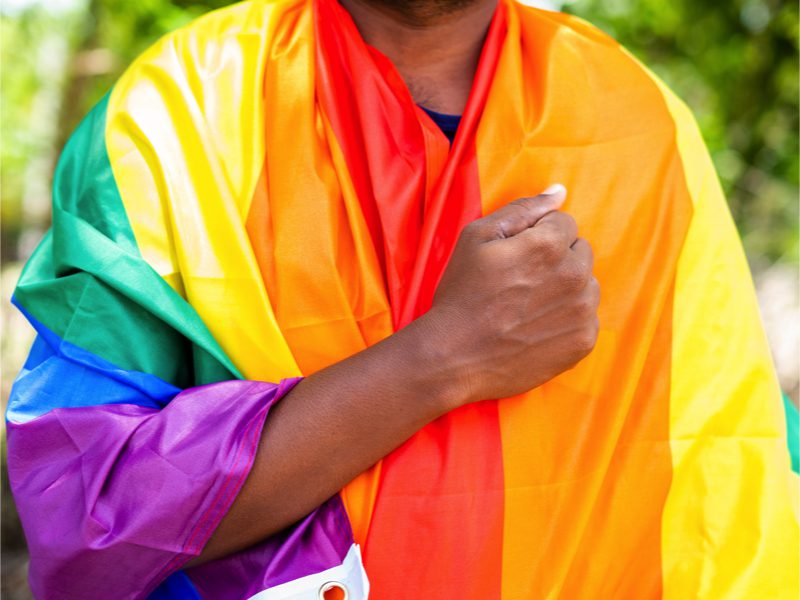 Photo of Man praying with clenched fist over heart while wearing rainbow flagcing during pride march