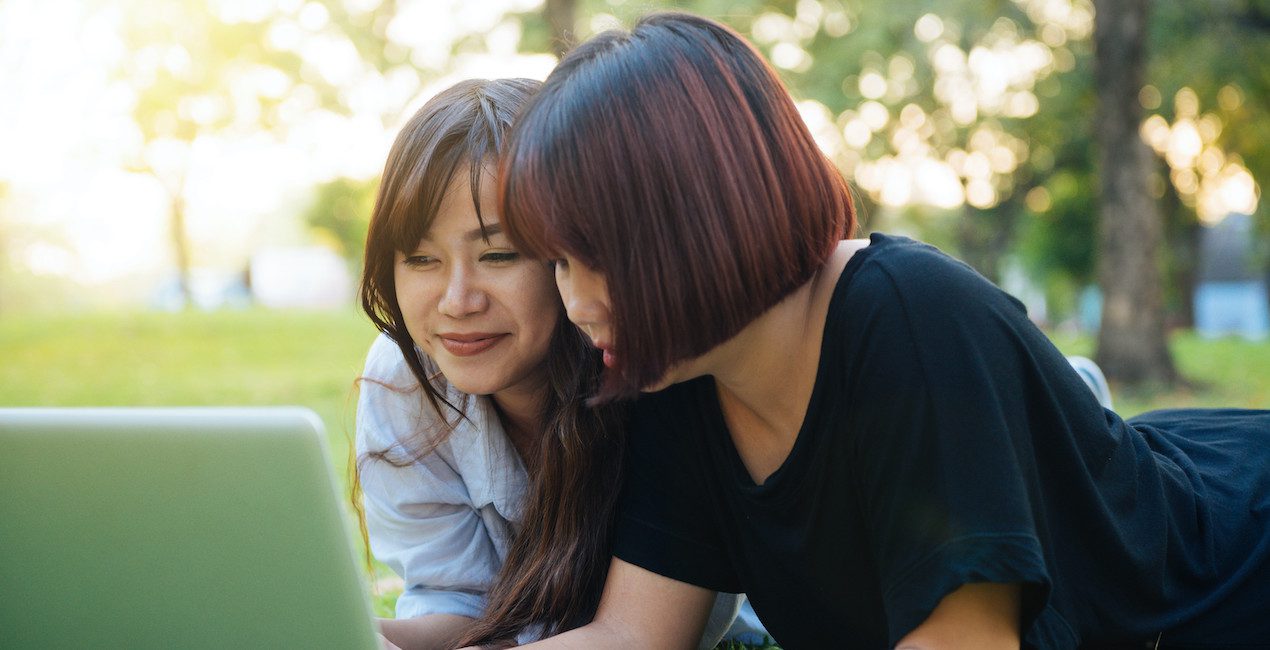 Two Asian women lying in the grass together looking at a laptop