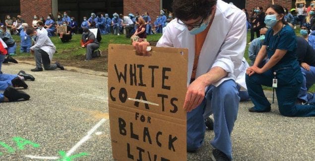 Healthcare worker kneeling with a sign that says "White coats for Black Lives Matter"