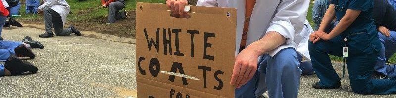 Healthcare worker kneeling with a sign that says "White coats for Black Lives Matter"