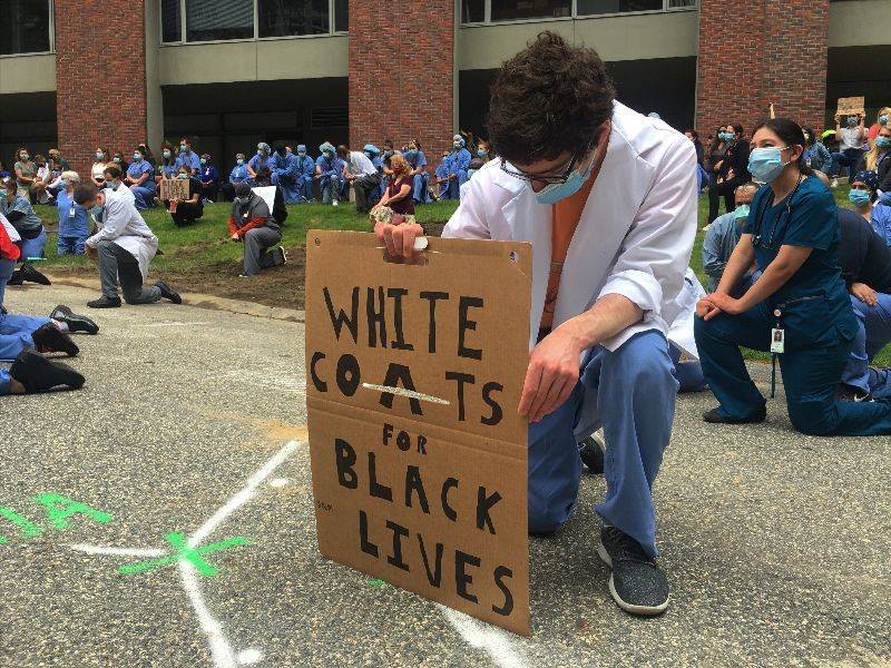 Healthcare worker kneeling with a sign that says "White coats for Black Lives Matter"