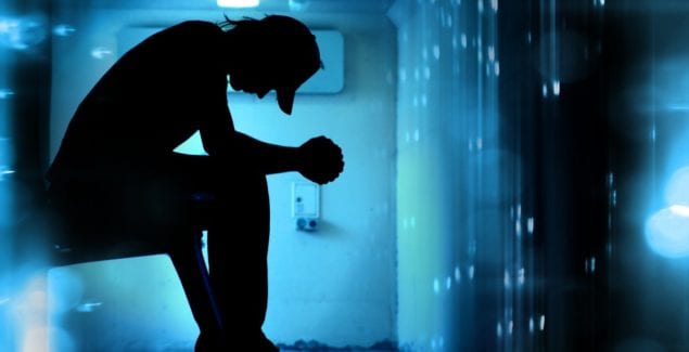 Young man sits on ledge in dark room with elbows on knees, looking down