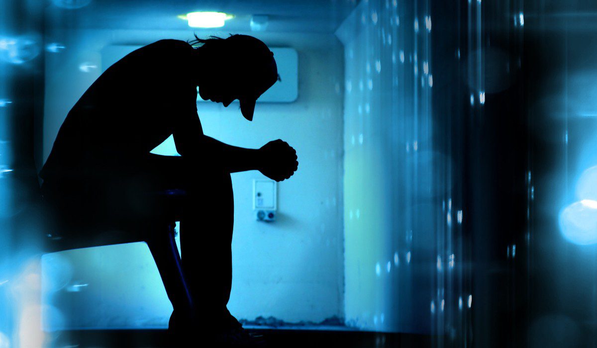 Young man sits on ledge in dark room with elbows on knees, looking down