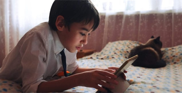 A young teen boy in a white shirt with a tie uses a tablet to communicate in video chat, in his bedroom, with a cat by his side.