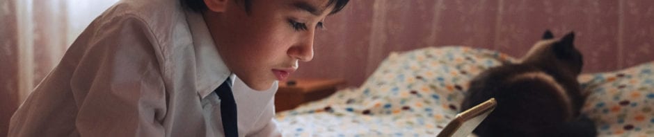 A young teen boy in a white shirt with a tie uses a tablet to communicate in video chat, in his bedroom, with a cat by his side.