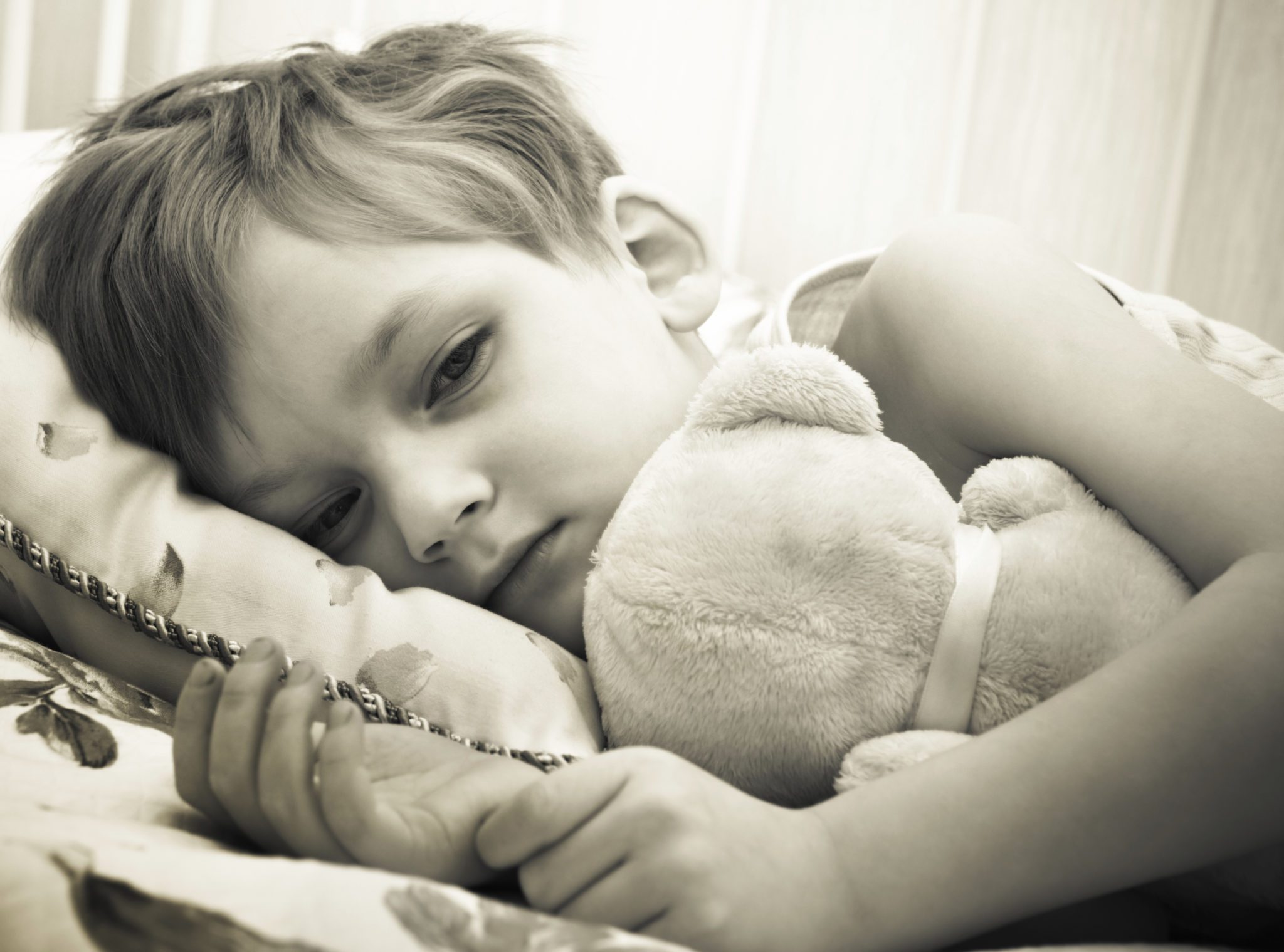 Sepia tone photo of young child cuddling their teddy in bed - measles, flu