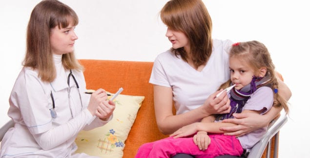 Female doctor sits on a couch next to mother holding sick young girl with thermometer in her mouth