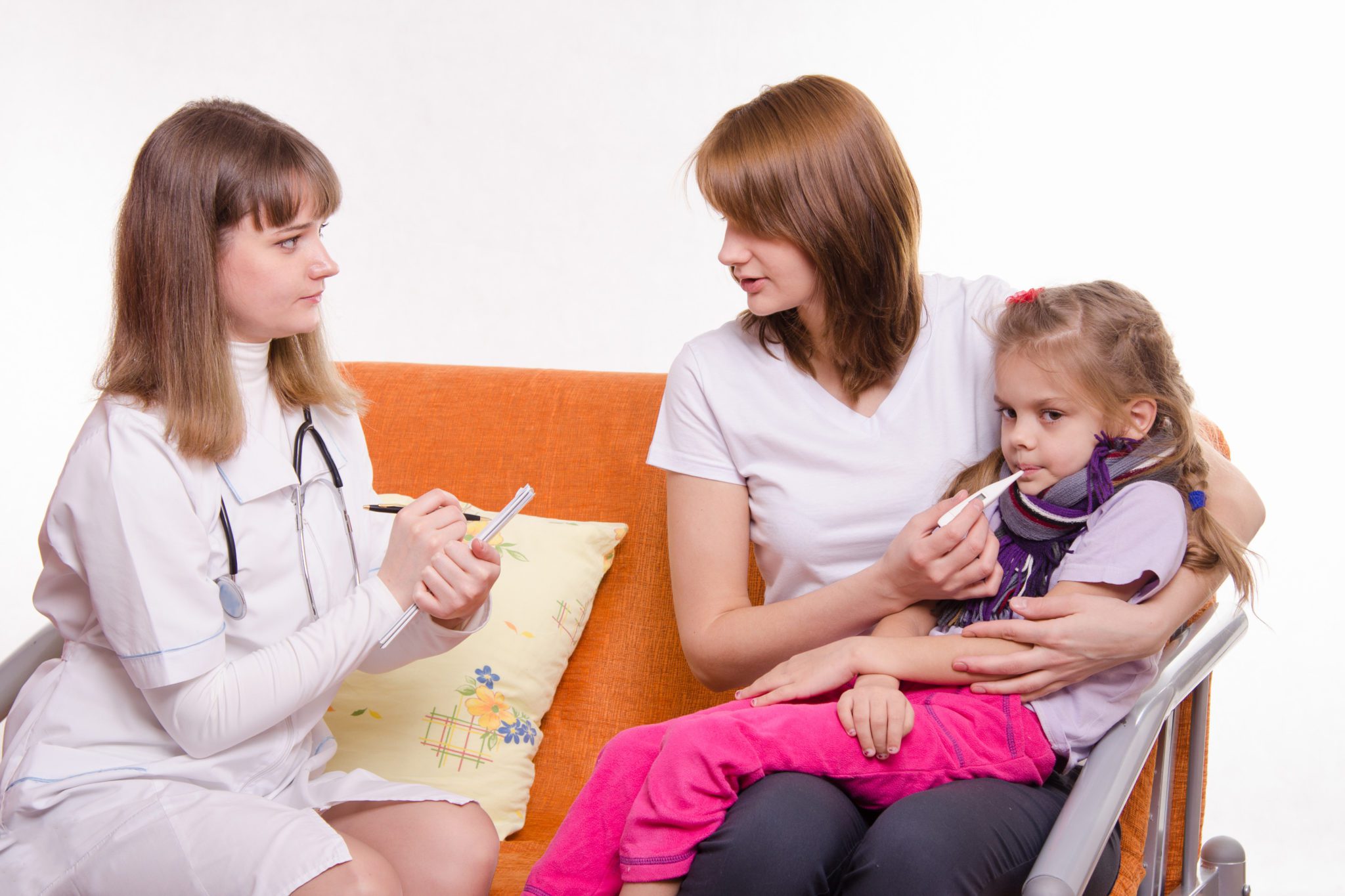 Female doctor sits on a couch next to mother holding sick young girl with thermometer in her mouth