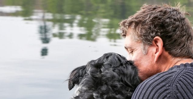 Behind view of man and dog sitting and hugging in front of lake