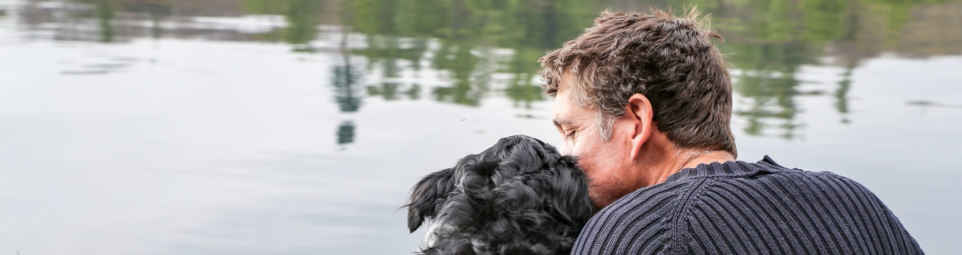 Behind view of man and dog sitting and hugging in front of lake
