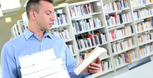 Middle-aged man reads back cover of book while holding stack of books in one arm
