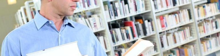 Middle-aged man reads back cover of book while holding stack of books in one arm