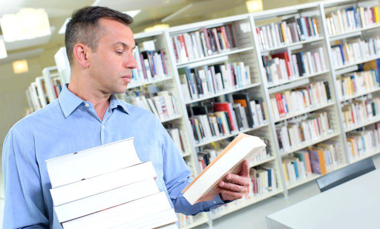 Middle-aged man reads back cover of book while holding stack of books in one arm