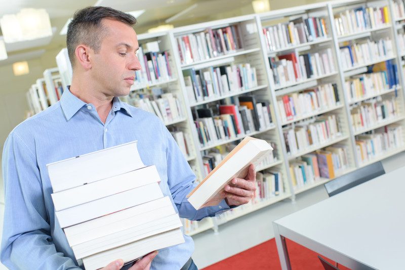 Middle-aged man reads back cover of book while holding stack of books in one arm