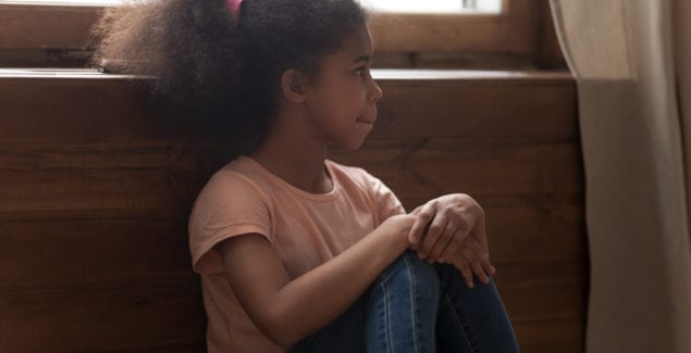 high functioning autism concept - Pensive african preschool or school age girl sitting on floor alone, leaned on wooden wall puts hands on laps looking away