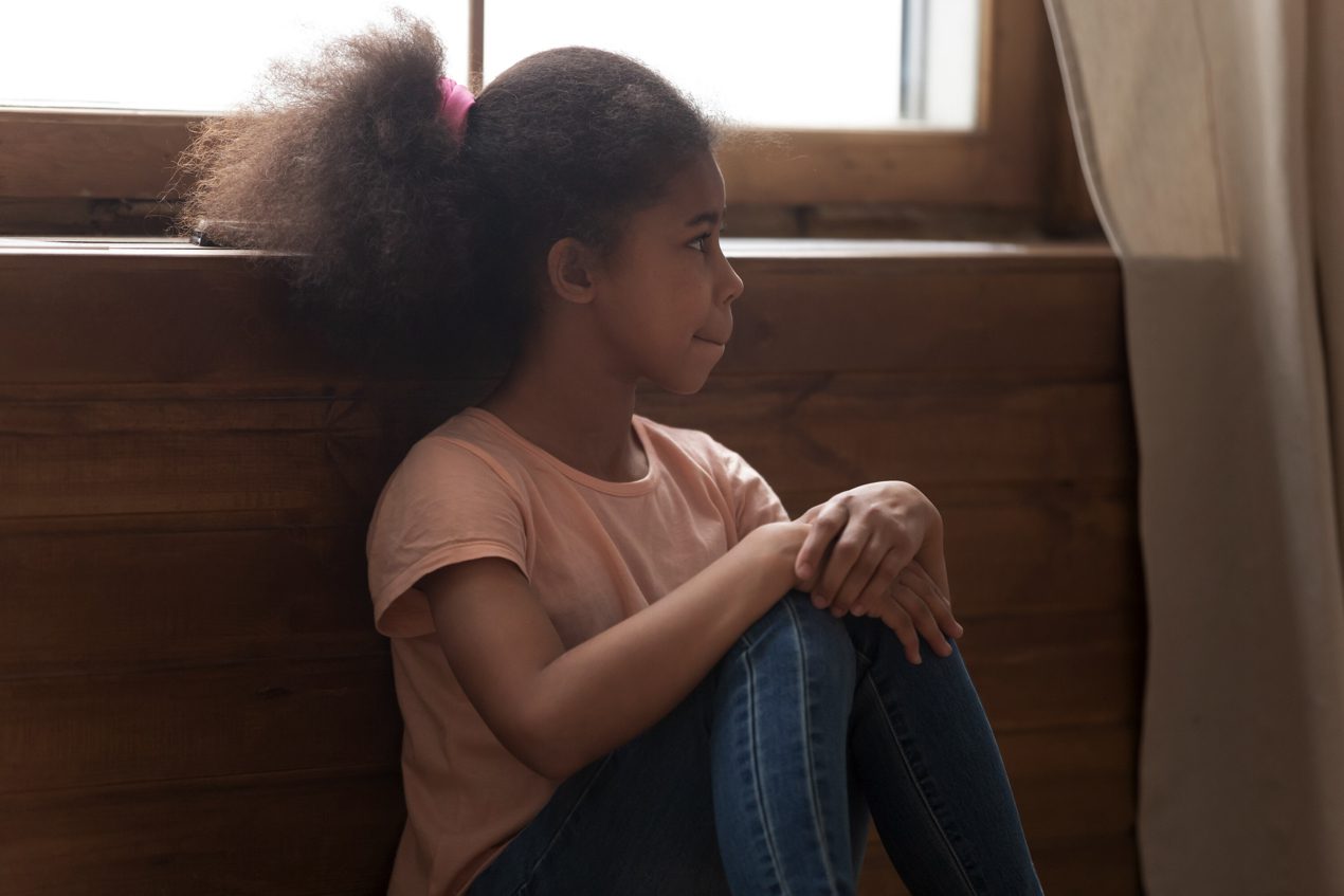 high functioning autism concept - Pensive african preschool or school age girl sitting on floor alone, leaned on wooden wall puts hands on laps looking away