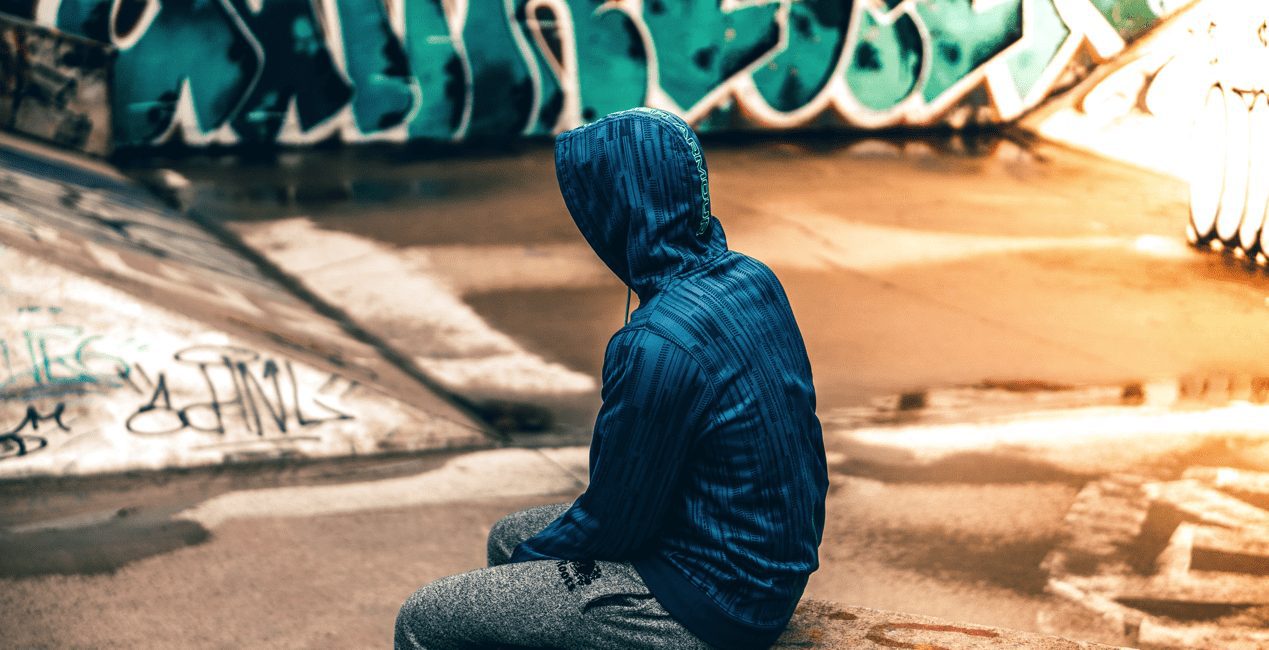 Teen sitting alone in front of graffiti wall with hood pulled up over head.