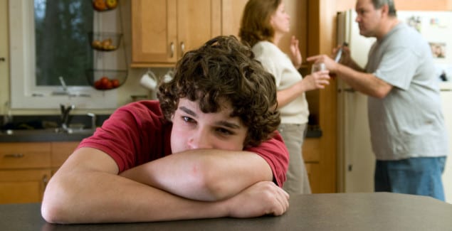 Divorce - adolescent leaning on table with head in arms, with fighting parents in the background