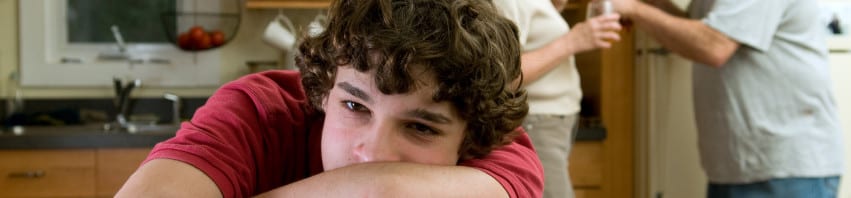 Divorce - adolescent leaning on table with head in arms, with fighting parents in the background