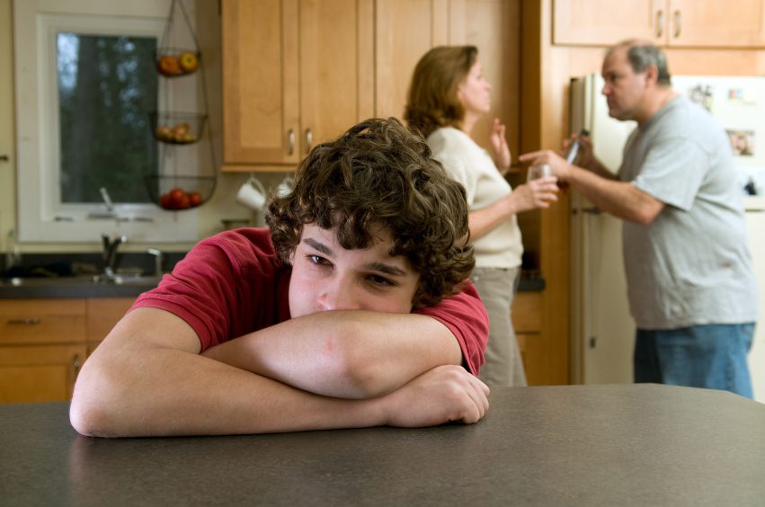 Divorce - adolescent leaning on table with head in arms, with fighting parents in the background